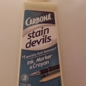 K&ouml;nnte beinhalten: Eine Flasche Carbona Stain Devils Fleckenentferner f&uuml;r Tinten-, Marker- und Buntstiftflecken. Die Flasche ist hellgelb mit wei&szlig;em Deckel und blauem Etikett mit Produktname und einer Grafik von Stiften und Bleistiften. Enth&auml;lt 50ml.