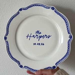 Puede incluir: Un plato de cerámica blanca con borde festoneado y una cenefa decorativa azul. El centro del plato está personalizado con el texto azul "the Harpers" en letra cursiva sobre la fecha "10.03.26" escrita en una fuente sencilla sin remates.