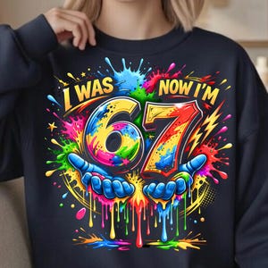 May include: Navy blue sweatshirt featuring the text "I WAS 67 NOW I'M" in a vibrant, multi-coloured design. The number 67 is the focal point, held by blue hands, with colourful splashes and lightning bolts surrounding it.