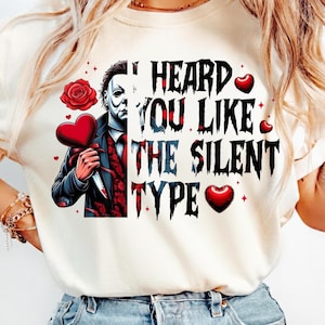 May include: A cream-colored t-shirt featuring a graphic of a masked character in a suit holding a heart-shaped object and a knife. The text reads, I HEARD YOU LIKE THE SILENT TYPE, accompanied by red rose and heart illustrations in a horror-themed style.