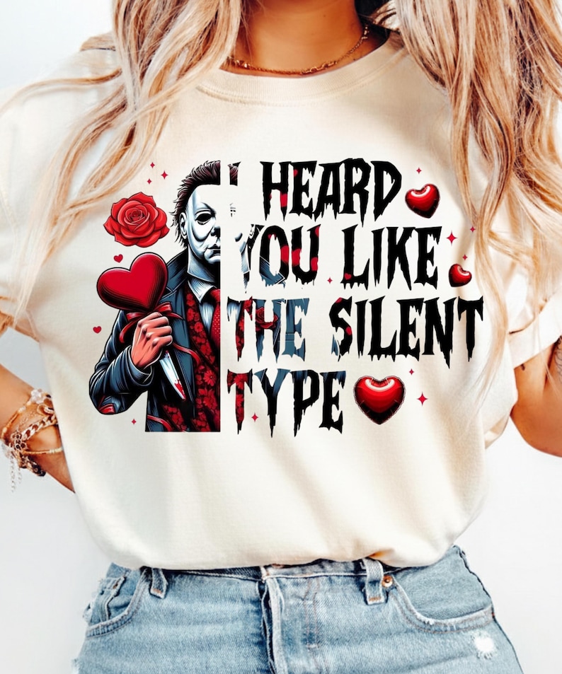 May include: A cream-colored t-shirt featuring a graphic of a masked character in a suit holding a heart-shaped object and a knife. The text reads, I HEARD YOU LIKE THE SILENT TYPE, accompanied by red rose and heart illustrations in a horror-themed style.