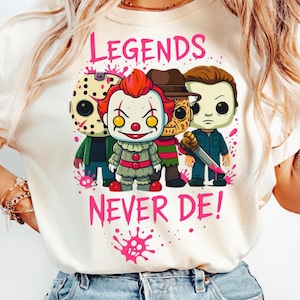 May include: A cream-colored t-shirt featuring a graphic of four horror movie characters in a stylized chibi design. Above and below the characters, the text reads "LEGENDS NEVER DE!" in pink, accompanied by pink paint splatter accents.