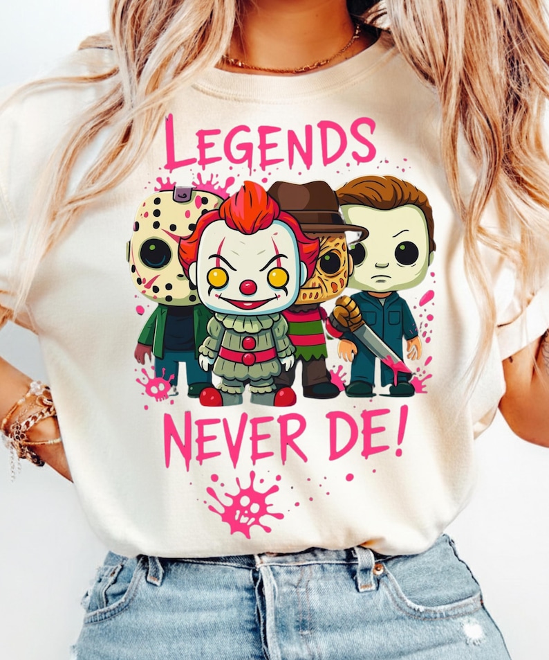 May include: A cream-colored t-shirt featuring a graphic of four horror movie characters in a stylized chibi design. Above and below the characters, the text reads "LEGENDS NEVER DE!" in pink, accompanied by pink paint splatter accents.