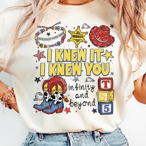 May include: A cream-colored t-shirt featuring a graphic print with a beaded bracelet, a yellow sheriff badge, a red cowboy hat, blue and white cow-print boots, and three stacked blocks labeled T, S, and 5. Text reads I KNEW IT I KNEW YOU and infinity and beyond.
