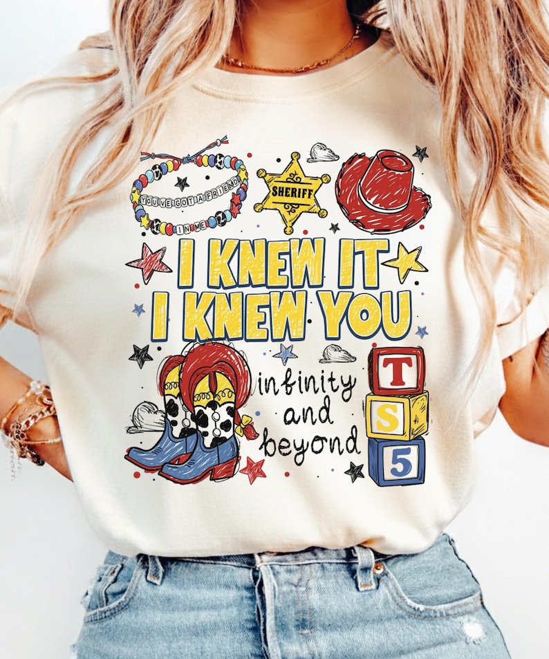 May include: A cream-colored t-shirt featuring a graphic print with a beaded bracelet, a yellow sheriff badge, a red cowboy hat, blue and white cow-print boots, and three stacked blocks labeled T, S, and 5. Text reads I KNEW IT I KNEW YOU and infinity and beyond.