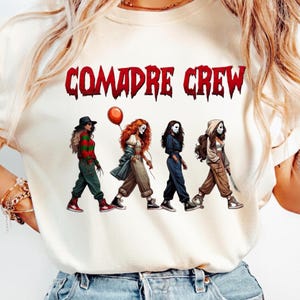 May include: A cream-colored t-shirt featuring the red text COMADRE CREW above a graphic of four figures dressed as horror movie characters. The figures are walking in a line, wearing various outfits including a striped sweater, a clown costume with a red balloon, a dark jumpsuit, and a hooded jacket with a mask.