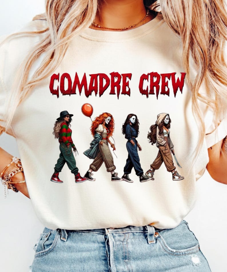 May include: A cream-colored t-shirt featuring the red text COMADRE CREW above a graphic of four figures dressed as horror movie characters. The figures are walking in a line, wearing various outfits including a striped sweater, a clown costume with a red balloon, a dark jumpsuit, and a hooded jacket with a mask.