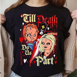 May include: A black t-shirt featuring a graphic print of two horror movie dolls, Chucky and Tiffany, separated by a large knife. The text Till Death Do Us Part is printed in a stylized font, accompanied by red hearts, blood drips, and a brick wall background.