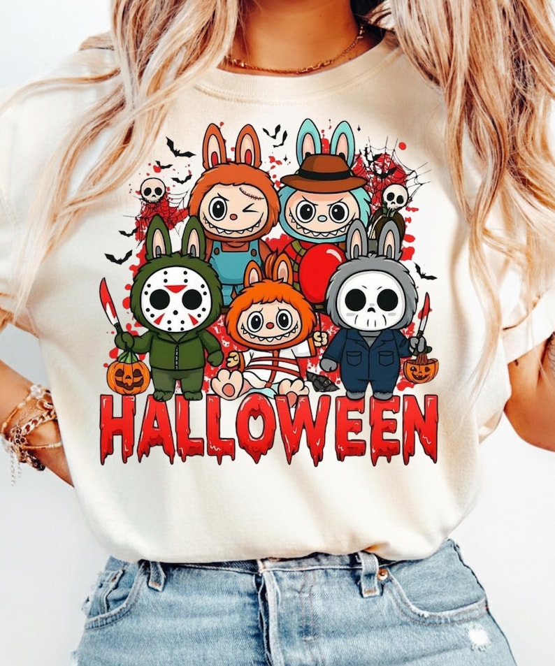 May include: A cream-colored t-shirt featuring a graphic print of five cartoon characters dressed in Halloween costumes, including masks and props, with the word HALLOWEEN in red, dripping, blood-like lettering at the bottom of the design.