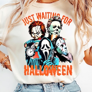 May include: A cream-colored t-shirt featuring a graphic print of four horror movie characters: Chucky, Michael Myers, Ghostface, and Jason Voorhees. The text "JUST WAITING FOR HALLOWEEN" is printed in orange above and below the characters.