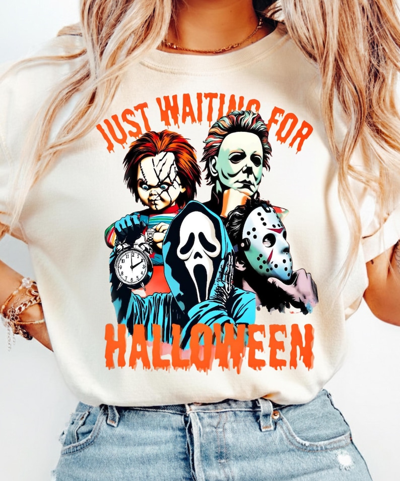 May include: A cream-colored t-shirt featuring a graphic print of four horror movie characters: Chucky, Michael Myers, Ghostface, and Jason Voorhees. The text "JUST WAITING FOR HALLOWEEN" is printed in orange above and below the characters.