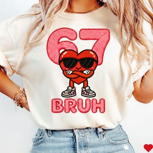 Puede incluir: Camiseta color crema con un personaje de corazón rojo con gafas de sol y zapatillas deportivas, el número 67 arriba y la palabra "BRUH" abajo. El diseño está bordado en un estilo texturizado.
