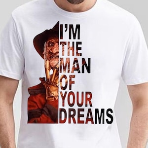 May include: A white short-sleeved t-shirt featuring a graphic of a character wearing a brown hat and a red and black striped sweater, with a clawed hand near the face. The text "I'M THE MAN OF YOUR DREAMS" is printed in black capital letters.