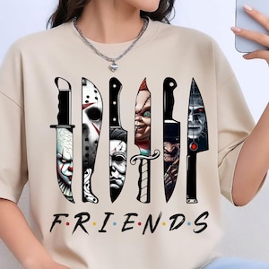 May include: A beige short-sleeved t-shirt featuring a graphic print of six knives, each containing a different horror movie character face. Below the knives, the word "FRIENDS" is printed in black with colorful dots between each letter.