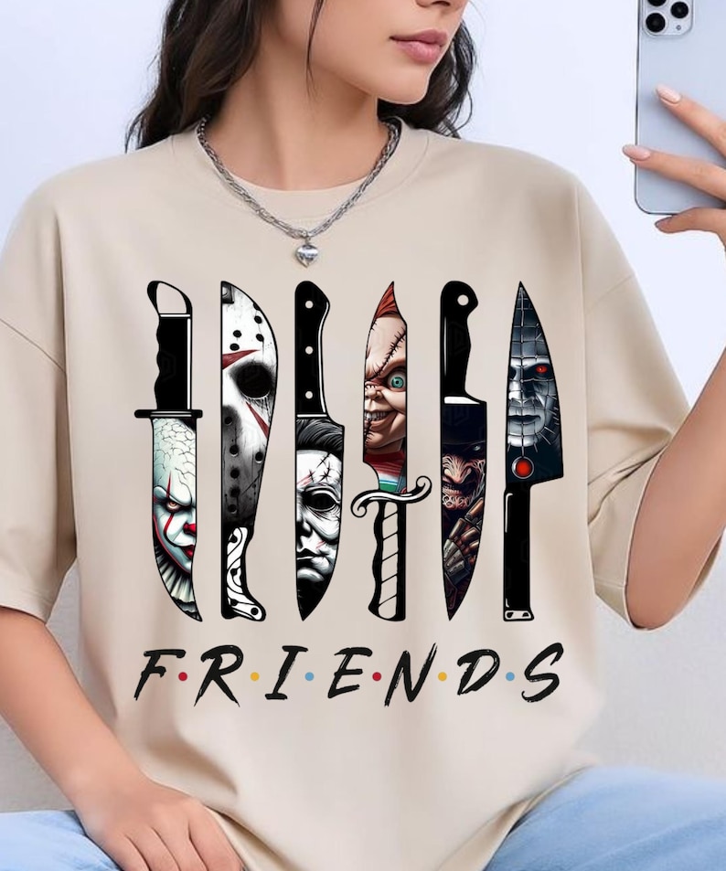 May include: A beige short-sleeved t-shirt featuring a graphic print of six knives, each containing a different horror movie character face. Below the knives, the word "FRIENDS" is printed in black with colorful dots between each letter.