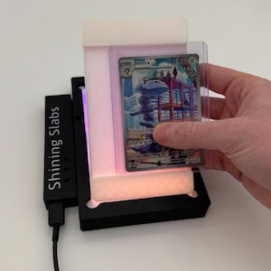 May include: A black device with the text "Shining Slabs" is connected to a power source. A person is holding a trading card in a clear protective sleeve inside a white and black illuminated display case. The card features a blue and gray illustration of a Steelix.