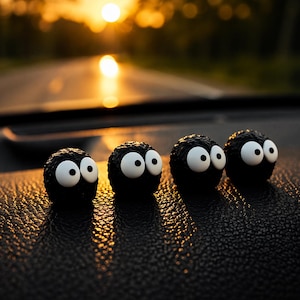 May include: Four small, black, spherical figures with large, white eyes are arranged in a row. The figures are on a textured, dark surface, with a blurred background of a road and a setting sun.