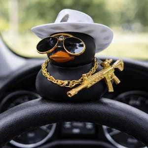 Pode incluir: Um pato de borracha preto usando um chapéu fedora branco, óculos de sol com armação dourada e uma corrente de ouro. O pato também tem um rifle de brinquedo dourado. O pato está sentado em um volante preto.
