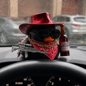 Könnte beinhalten: Eine schwarze Quietscheente mit einem metallisch roten Cowboyhut, schwarzer Sonnenbrille, rotem Paisley-Bandana und einer goldenen Kette. Sie hält eine Miniatur-Budweiser-Bierflasche und ein silberfarbenes Spielzeuggewehr auf einem Armaturenbrett.