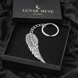 Puede incluir: Un llavero plateado en forma de ala, con cadena y anilla, presentado en una caja negra. La tapa de la caja muestra el texto "LUNAR MUSE STORE" y un logotipo de media luna. El llavero presenta texturas detalladas en forma de plumas.