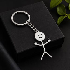 Peut inclure: Un porte-clés en métal argenté composé d'un anneau circulaire relié à une chaîne et d'un pendentif en forme de bonhomme allumette. Le bonhomme a une tête ronde avec un visage souriant et les bras levés, chaque main faisant un doigt d'honneur.
