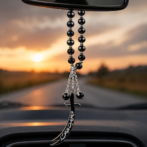 May include: Black beaded car charm hanging from a rearview mirror. The charm features a black sword pendant with Arabic script and two black beads at the bottom. The background shows a sunset over a road.