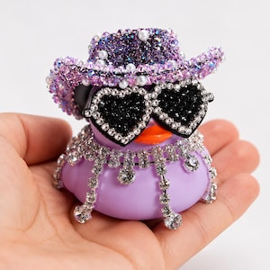 May include: A purple rubber duck figurine adorned with a purple rhinestone cowboy hat, black heart-shaped sunglasses with rhinestone frames, and a dangling silver-toned rhinestone necklace draped around its neck with an orange beak.