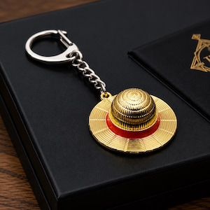 Może przedstawiać: Metalowy brelok do kluczy z zawieszką w kształcie miniaturowego słomkowego kapelusza o złotym, teksturowanym wykończeniu i z żywą czerwoną wstążką wokół główki. Zawieszka jest przymocowana do srebrnego łańcuszka i okrągłego kółka na klucze.
