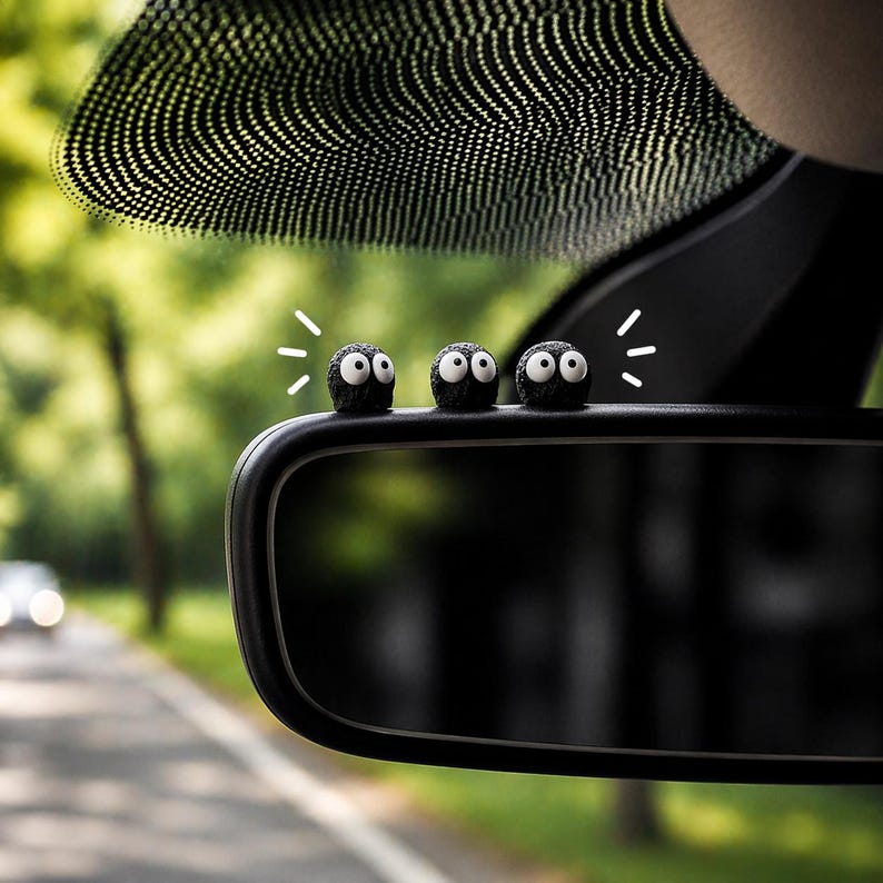 May include: Three small, black, round car accessories with white eyes are perched on a car's rearview mirror. The accessories have a fuzzy texture and are positioned in a row. The background shows a blurred view of a road and trees.