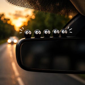 May include: A row of five black, round car accessories with white eyes and a surprised expression, attached to a car's rearview mirror. The background shows a road with a car and a sunset.
