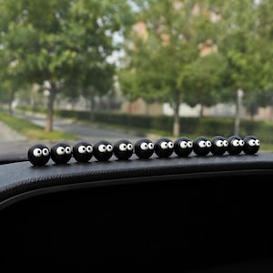 May include: A row of black, spherical decorative car accessories with white eyes and a smiling expression. The accessories are lined up on a black dashboard, with a blurred background of trees and a street.