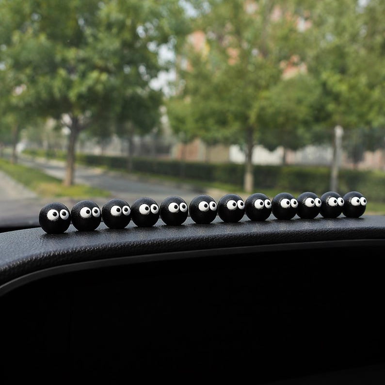 May include: A row of black, spherical decorative car accessories with white eyes and a smiling expression. The accessories are lined up on a black dashboard, with a blurred background of trees and a street.