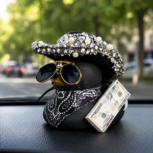 Könnte beinhalten: Eine schwarze Gummi-Ente mit einem schwarzen Cowboyhut, der mit silbernen Strasssteinen und weißen Perlen besetzt ist, einer schwarzen Sonnenbrille mit goldenem Rahmen, einem schwarz-weißen Paisley-Bandana und einem Bündel Geldscheine.
