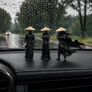 Può includere: Tre piccole figure di samurai neri con cappelli di paglia sono esposte su una superficie scura. Una figura tiene una spada, un'altra ha le braccia incrociate e la terza ha un mantello fluente. Sullo sfondo si vedono una strada bagnata e degli alberi.