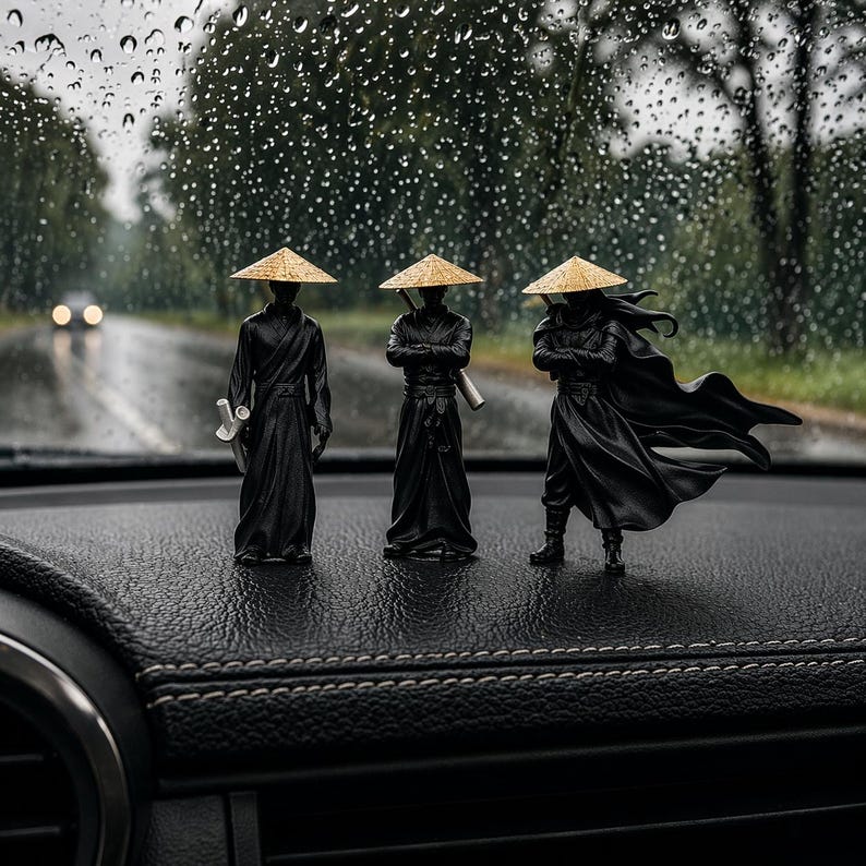 May include: Three miniature black samurai figures with straw hats stand on a dark surface. One figure holds a sword, another has crossed arms, and the third has a flowing cape. The background shows a wet road and trees.