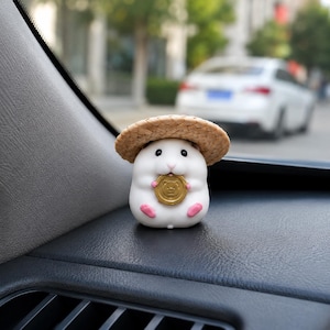 May include: A small white hamster figurine wearing a tan woven straw hat. The hamster has black eyes, a pink nose, and pink feet. It is holding a round gold-coloured coin featuring an embossed bear face design in its front paws.