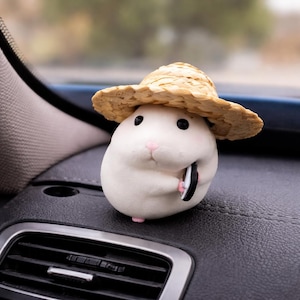 May include: A white, round figurine of a hamster wearing a straw hat. The hamster has black eyes, a pink nose, and is holding a small, black and white object. The figurine is placed on a black dashboard.