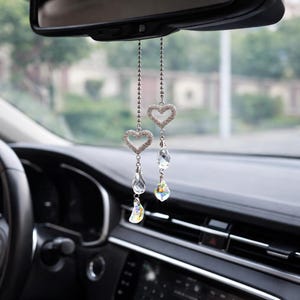 Könnte beinhalten: Zwei Auto-Rückspiegel-Anhänger mit herzförmigen Anhängern. Jeder Anhänger hat eine silberne Kette, ein herzförmiges Strass-Detail und einen klaren Kristalltropfen. Die Anhänger hängen vor dem Armaturenbrett und dem Rückspiegel eines Autos.
