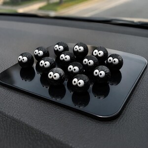 May include: A collection of black, spherical car dashboard decorations with large white eyes, arranged on a black rectangular pad. The decorations have a textured surface and are reflected on the pad's glossy finish.