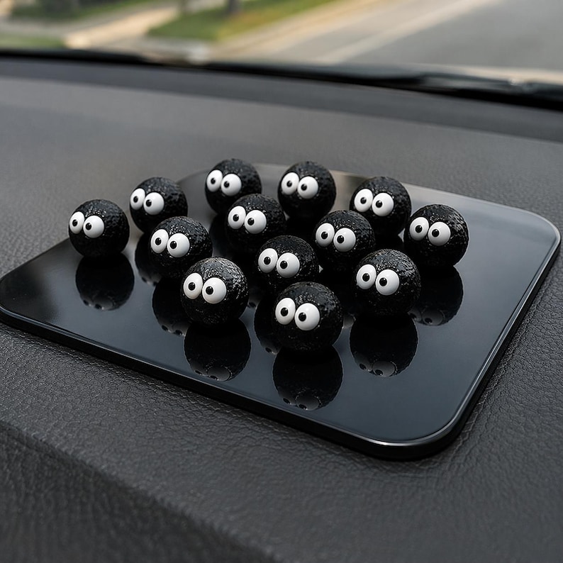 May include: A collection of black, spherical car dashboard decorations with large white eyes, arranged on a black rectangular pad. The decorations have a textured surface and are reflected on the pad's glossy finish.