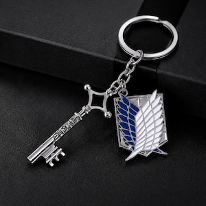 Puede incluir: Un llavero de metal plateado que presenta un dije de llave decorativo y un colgante en forma de escudo. El colgante muestra un emblema de alas en azul y blanco. La llave tiene un diseño de mango intrincado y un paletón estándar.