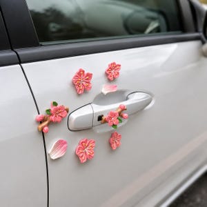 May include: A set of pink cherry blossom flower decorations attached to the side of a white car door. The set includes individual blossoms, small branches with buds and leaves, and detached petals arranged around the silver door handle.