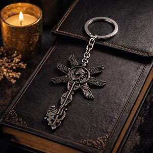 May include: A silver key chain with a detailed key design, featuring a skull and wing motif. The key chain rests on a dark leather-bound book. A lit candle in a gold holder is in the background.