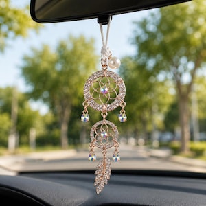 Puede incluir: Un adorno para coche en oro rosa con un diseño de atrapasueños. El adorno tiene dos marcos circulares con un patrón floral, adornados con cristales transparentes y detalles iridiscentes. Una perla y una pluma con incrustaciones de cristal completan el diseño, suspendidos de un cordón blanco.
