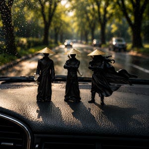 May include: Three black figurines of samurai warriors wearing conical straw hats with gold trim. The figures are posed on a dark surface, with a blurred road and trees in the background. The figures are in various stances, some holding weapons.