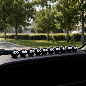 May include: A row of eleven black, spherical car dashboard decorations with white eyes. The decorations are lined up on a black dashboard, with a blurred background of green trees and a road.