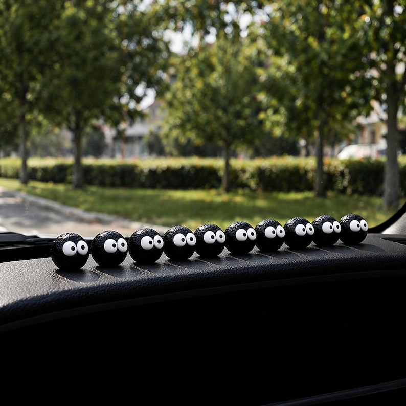 May include: A row of eleven black, spherical car dashboard decorations with white eyes. The decorations are lined up on a black dashboard, with a blurred background of green trees and a road.