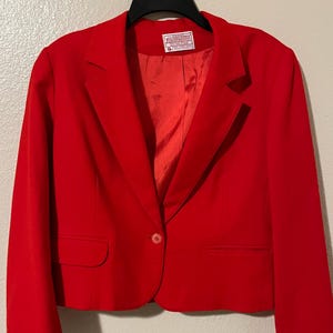 May include: A bright red blazer with a single-button closure and two front pockets. The blazer is hanging on a black hanger. The interior lining is a matching red color.