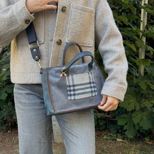 Könnte beinhalten: Graue Wildlederhandtasche mit einem karierten Aufnäher und blauen Lederbesatz. Die Tasche hat einen oberen Griff und einen abnehmbaren marineblauen Schulterriemen. Ein kleiner Anhänger hängt am Griff. Die Tasche wird über grauen Jeans und einer grauen Jacke getragen.