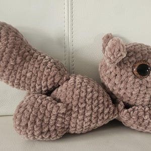 Puede incluir: Una ardilla de peluche hecha a mano, de color malva apagado. El peluche tiene una cola grande y esponjosa, orejas pequeñas y una cara detallada con ojos grandes y brillantes. La ardilla está hecha de un hilo suave y texturizado.